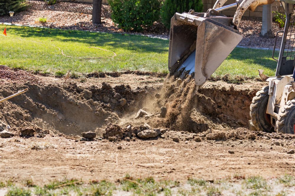 Excavator digging a trench in a yard, with soil being loaded into the bucket.